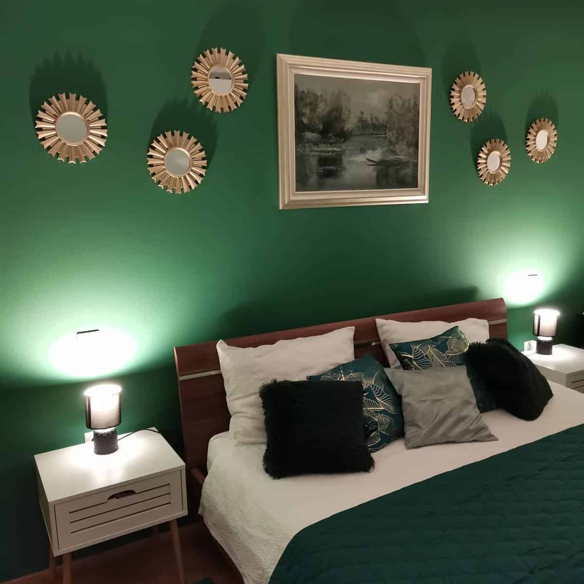 Cozy bedroom with green walls, mirror decor, framed painting, and a bed with green quilt and cushions