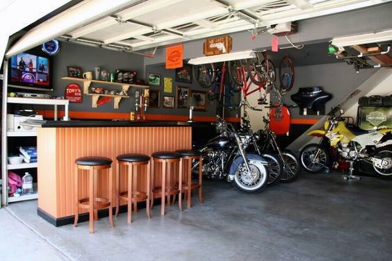 Garage bar with orange counter, wooden stools, hanging bikes, and parked motorcycles.