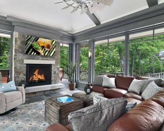 The sunroom features a stone fireplace, large leather sectional sofa, and floor-to-ceiling windows for a bright, modern look.