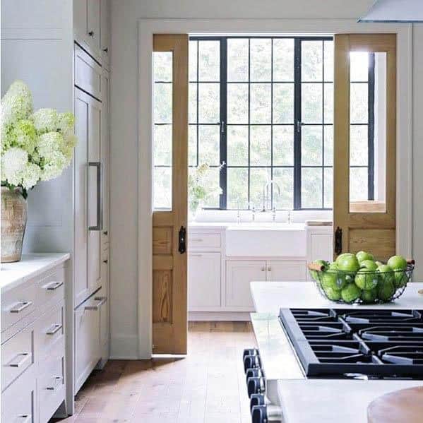 Natural wood pocket doors with glass panels open to a bright farmhouse kitchen featuring a large window, white cabinetry, and fresh decor