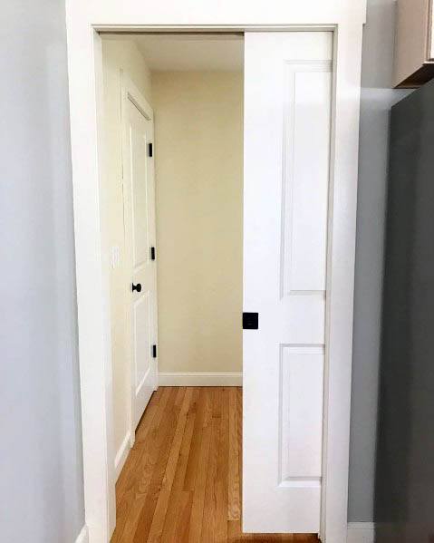 Simple white pocket door with black hardware opens to a clean, minimal hallway featuring light wood flooring and neutral walls