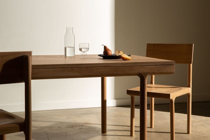 Minimalist dining setup with wooden table, chairs, and a carafe with fruit centerpiece.