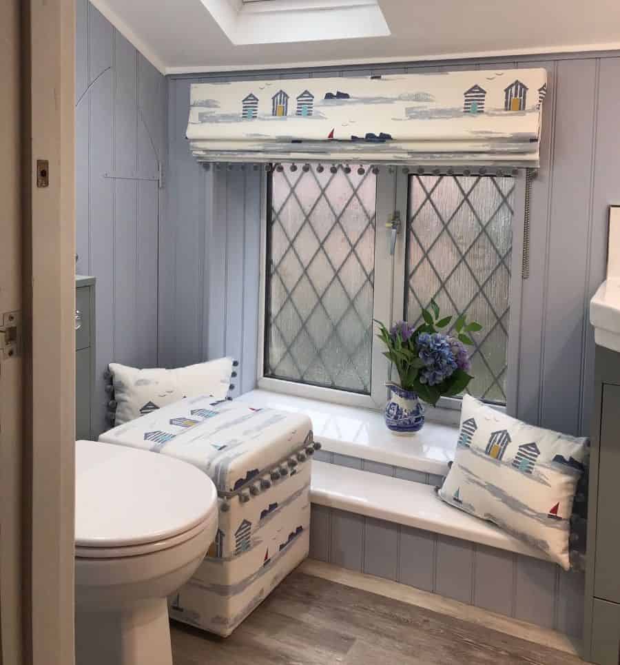 Coastal-themed bathroom with skylight, frosted windows, and decorative cushions by a seat with flowers