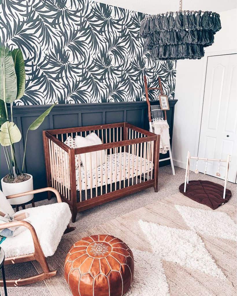 Boho nursery with dark accent wall, tropical wallpaper, wooden crib, and leather pouf.