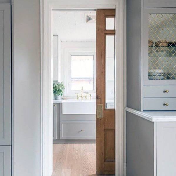 Natural wood pocket door with glass panels opens to a bright kitchen pantry featuring a farmhouse sink, gold fixtures, and soft cabinetry