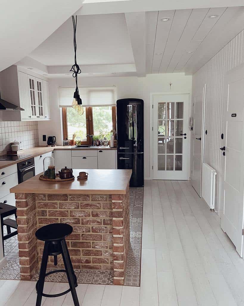 Cozy kitchen with brick island, black fridge, white cabinets, and natural light from windows.