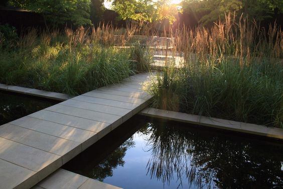 Modern stone bridge crossing a reflective pond, surrounded by tall grasses and natural landscaping.