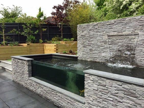 Raised stone pond with a glass window, featuring a waterfall and surrounded by modern garden landscaping.