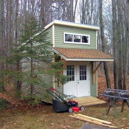 Two-story shed with a small canopy, double doors, and a lawnmower in a forested backyard.