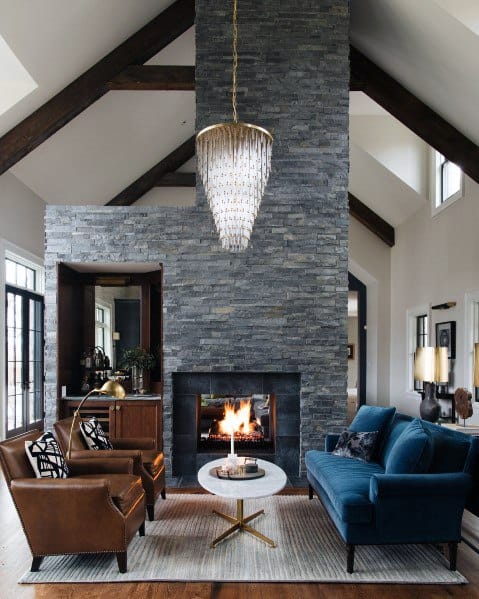 Cozy living room with a stone fireplace, chandelier, brown chairs, blue sofa, and a round coffee table