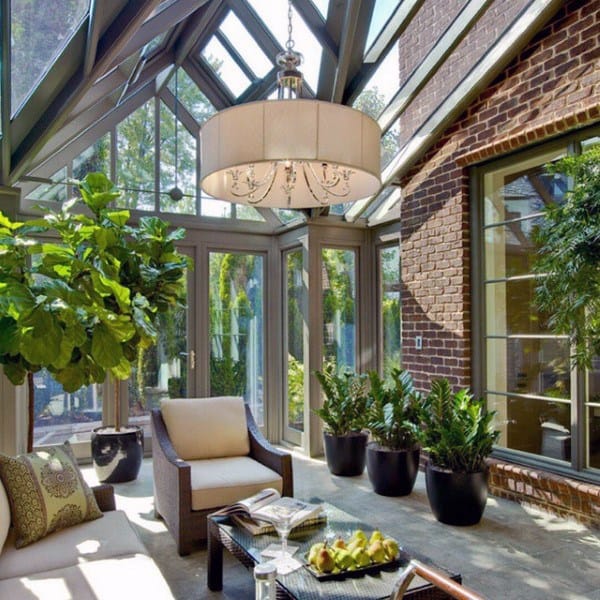 The sunroom features glass walls, a modern chandelier, lush potted plants, and cozy seating against a brick wall.