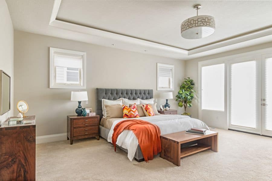 Spacious beige bedroom with wooden furniture, tufted bed, and orange accent pillows.