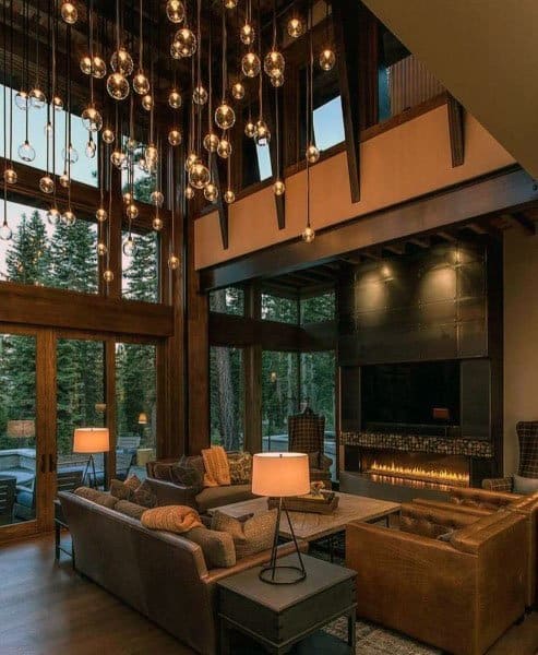 Cozy living room with high ceilings, hanging lights, large windows, and a modern fireplace overlooking the forest