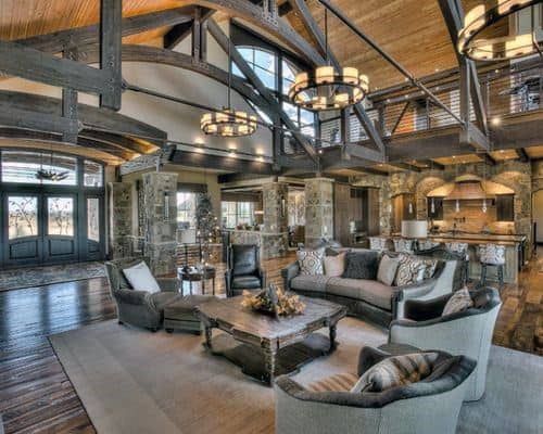 A spacious rustic living room with exposed beams, large windows, comfortable seating, and a connection to an open kitchen area, creating an airy and inviting space