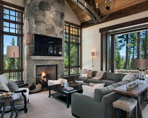 A rustic living room with a stone fireplace, modern furniture, large windows offering scenic views, and cozy decor, creating a warm and inviting atmosphere