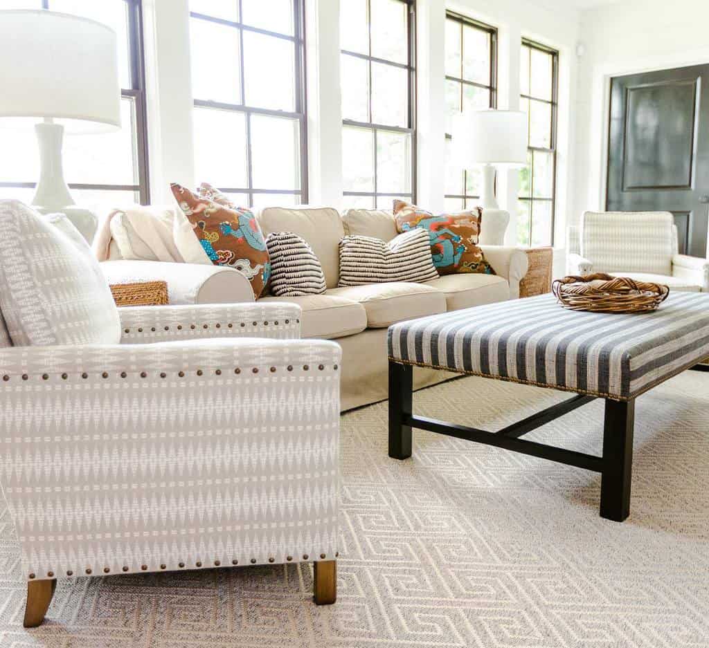 Bright sunroom with beige sofa, patterned armchairs, striped ottoman, and cozy decor.