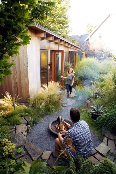 Wooden shed with large glass doors, lush garden path, and cozy fire pit seating area.