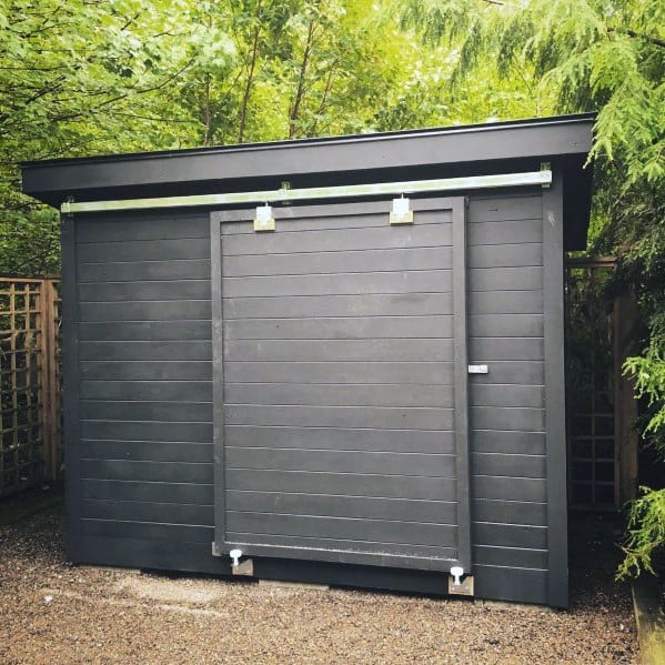 Sleek black box-style shed with a sliding barn door, set in a lush garden.