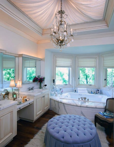 Elegant bathroom with chandelier, large tub, vanity, and blue tufted ottoman; draped ceiling and windows with garden view