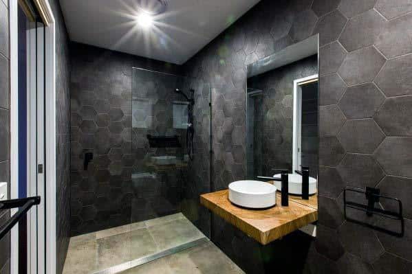 Dark modern bathroom with hexagon tiles, floating wooden vanity, and glass shower enclosure.