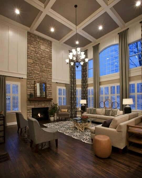 Spacious living room with high ceiling, stone fireplace, large windows, brown sectional sofa, and modern chandelier