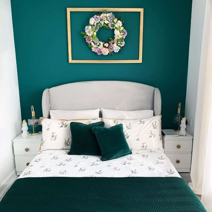 Small green bedroom with floral wreath decor, velvet pillows, and white bedside tables.