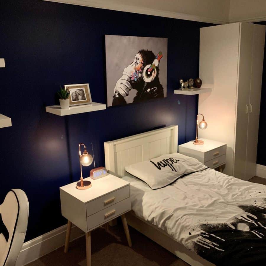 Modern kids' bedroom with dark blue walls, wall shelves, art, and white furnishings.