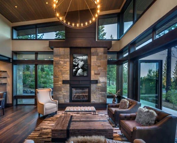 A cozy living room with a stone fireplace, plush chairs, a chandelier, and large windows overlooking a forest