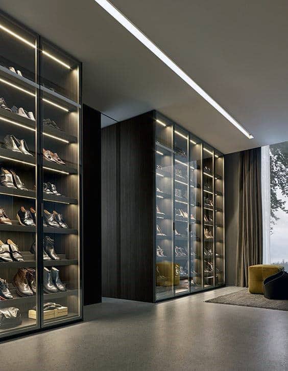 A modern shoe closet with glass doors displaying rows of neatly arranged shoes, next to a large window with curtains