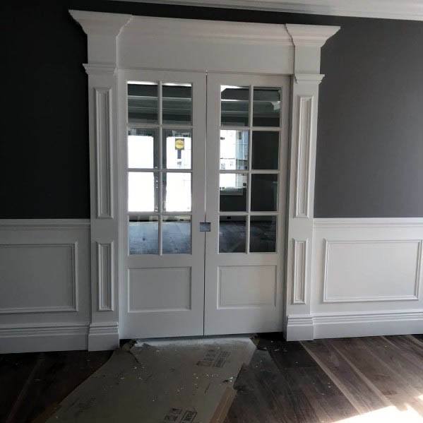 Elegant white double pocket doors with glass panels and intricate molding open to a bright dining space, featuring classic wainscoting