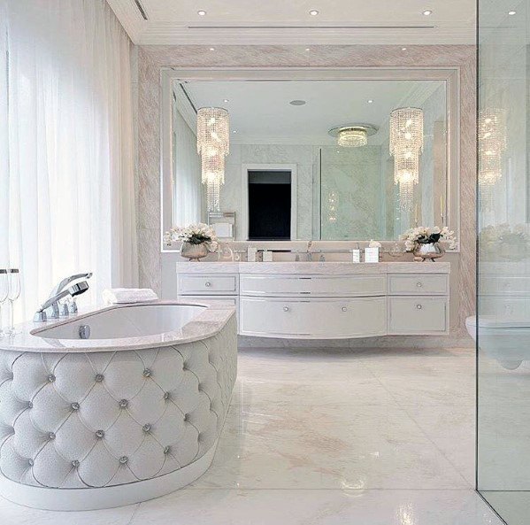 Luxurious white bathroom with a tufted bathtub, large mirror, and elegant chandeliers