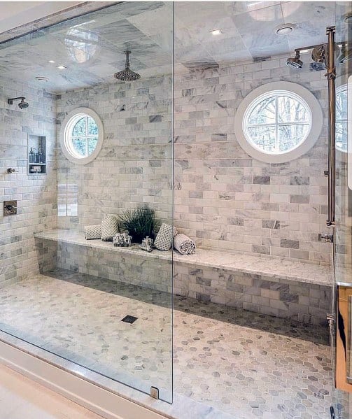 Spacious shower with marble tile, built-in bench, circular windows, and rainfall showerhead.