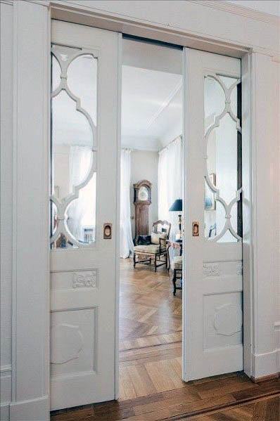 Ornate white pocket doors with decorative glass panels open to a bright, elegant room featuring parquet floors and vintage decor