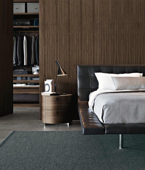 Modern bedroom with a leather bed, gray bedding, round nightstand, lamp, and open closet with hanging clothes