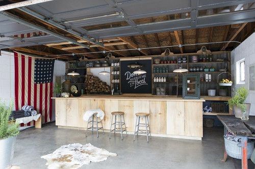 Rustic garage bar with wooden counter, open shelving, American flag decor, and industrial lighting.