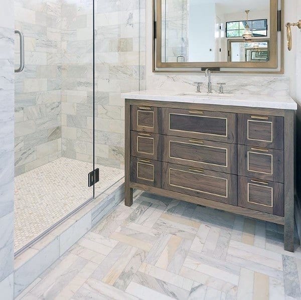 Bathroom with herringbone marble floor tiles, wood vanity with brass accents, and glass shower enclosure.