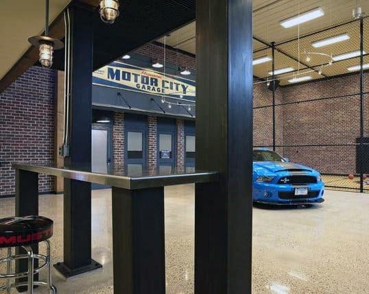 Industrial-themed garage bar with exposed brick walls, modern bar table, and a polished concrete floor featuring a classic car display.