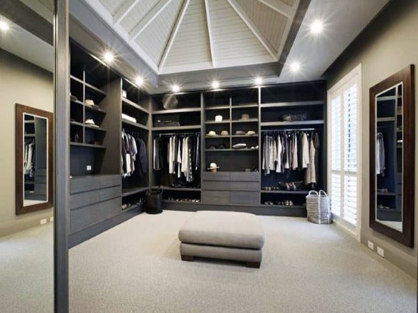 Spacious walk-in closet with built-in shelves, hanging clothes, and a central ottoman under a vaulted ceiling