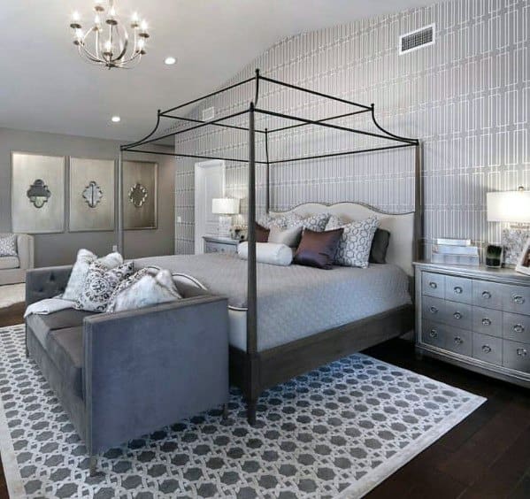 Elegant bedroom with patterned wallpaper, canopy bed, gray ottoman, and silver nightstands.