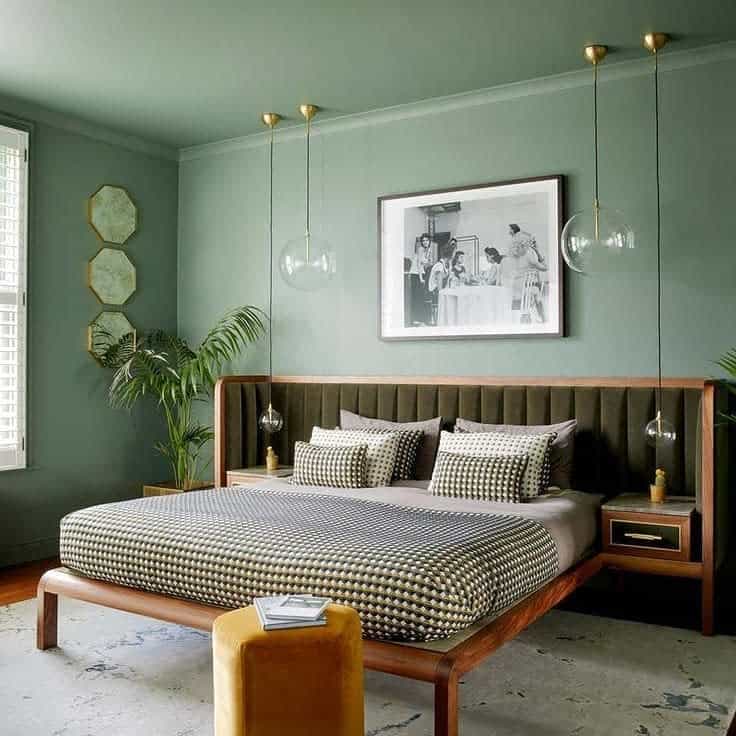 Stylish bedroom with green walls, patterned bedding, hanging lights, plants, and framed art above the bed