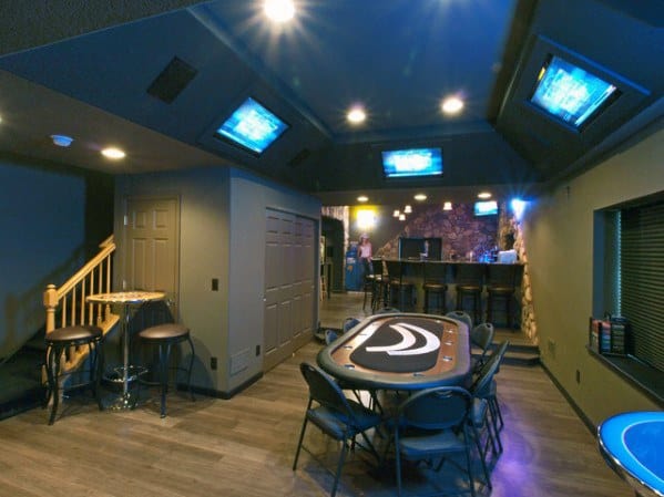 Man cave with poker table, multiple TV screens, bar area, and dim ambient lighting.