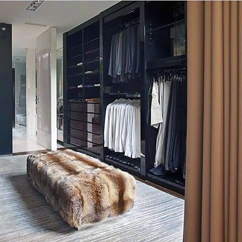 Spacious walk-in closet with black shelves, hanging clothes, and a fur bench on a rug, with curtains partially visible