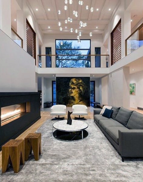 Modern living room with high ceiling, gray sofa, round table, fireplace, and hanging lights. Large windows in background