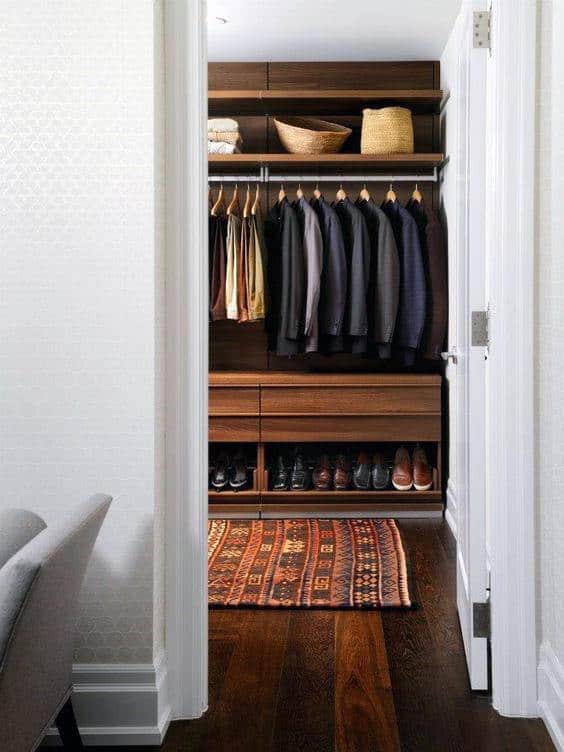 A neatly organized walk-in closet with suits, shirts, shoes, and baskets, featuring wooden shelves and a patterned rug