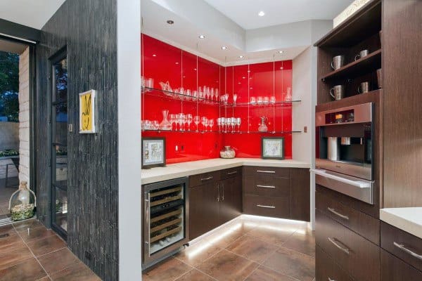 Modern mini bar with red glass backsplash, dark cabinets, wine fridge, and illuminated shelving.