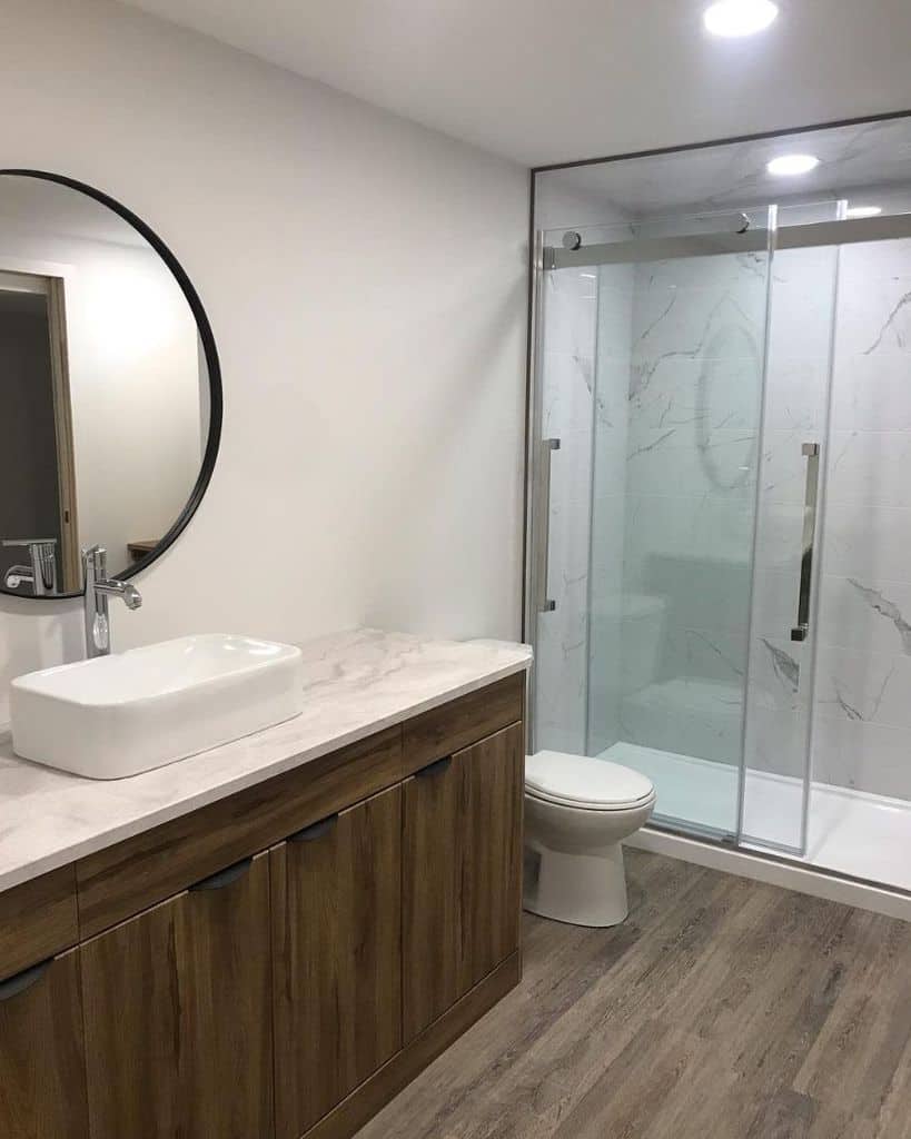 Minimalist basement bathroom with wood vanity, round mirror, and glass-enclosed shower.