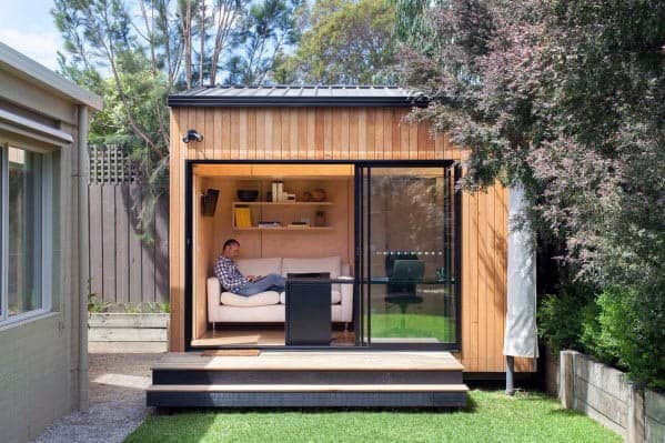 Small wooden box-style shed with sliding glass doors and cozy interior, set in a landscaped backyard.