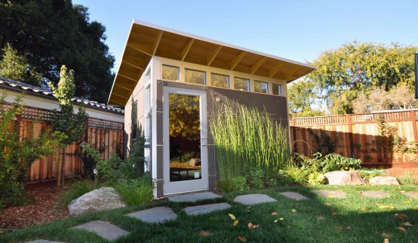 Modern greenhouse shed with large windows and door, surrounded by plants and greenery in a fenced yard.