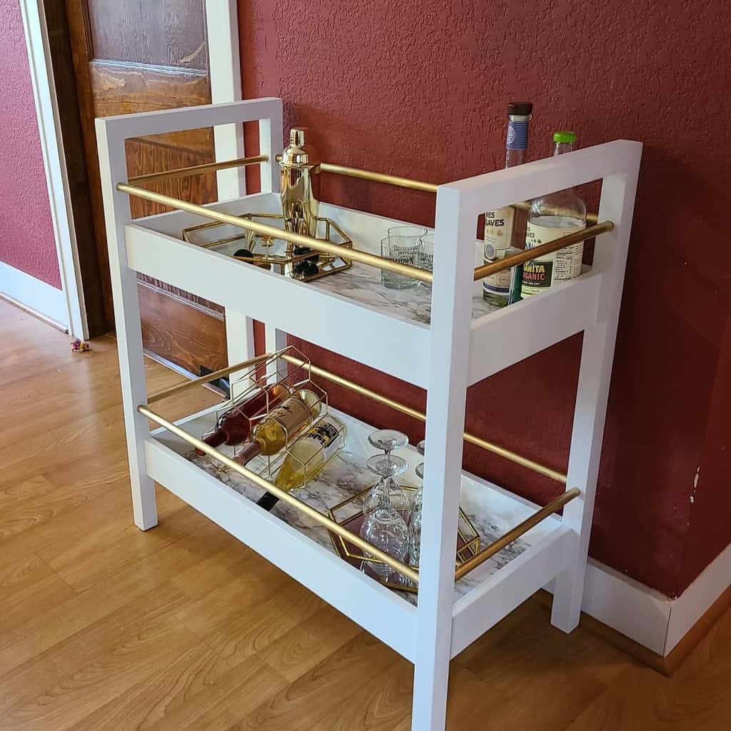 White bar cart with gold accents holding liquor bottles, glassware, and cocktail shaker.