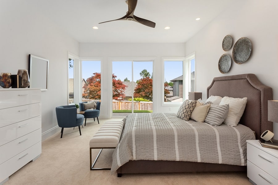 Modern bedroom with a gray upholstered bed, large windows, and a sleek ceiling fan.